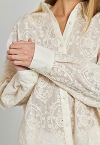 Bestickte Bluse in Creme, mit zarten Blumenmustern und einer durchgehenden Knopfleiste. Weicher Stoff mit strukturiertem Finish und Kragen-Design.