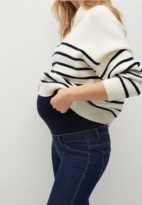 Mujer embarazada levantando un suéter de rayas para mostrar unos jeans de maternidad oscuros sobre su barriga, de pie contra un fondo sencillo.