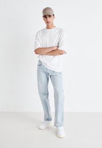 White oversized t-shirt, light blue straight-leg jeans, and white sneakers. Model wearing a light beige cap, arms crossed, plain background.
