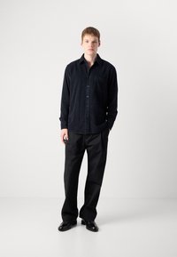 Camisa de pana negra con diseño de botones, bolsillo en el pecho y mangas largas, combinada con pantalones negros a medida y zapatos pulidos.