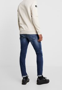 Sweat-shirt beige clair à manches longues avec poignets côtelés, associé à un jean bleu foncé slim fit. Chaussures de sport noires avec logo visible.