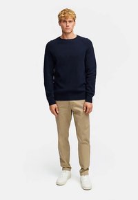 Mörkblå stickad tröja med ribbade muddar och fåll, tillsammans med beige byxor och vita sneakers, stående mot en enfärgad bakgrund.