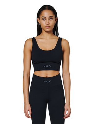 Kobieta z plecionymi włosami, ubrana w czarny sportowy biustonosz i legginsy z wysokim stanem, oba z logo "HALO" z przodu.