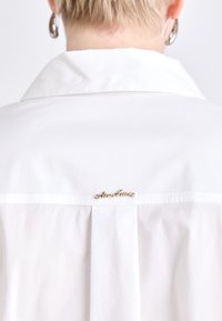 Blusa blanca con cuello extendido, que presenta un pliegue en la parte posterior y un acento de logo en escritura dorada cerca del escote. Tela suave y ligera.