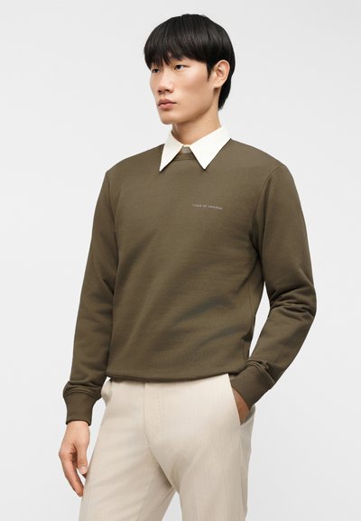 Φούτερ σε ελαιί χρώμα με στρογγυλή λαιμόκοψη, που φέρει ένα λευκό πουκάμισο με γιακά από κάτω. Διακριτικό λογότυπο τυπωμένο στο στήθος σε ανοιχτό γκρι.