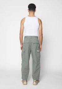 Zielone spodnie cargo z wieloma kieszeniami i naszywką z logo, wykonane z wytrzymałej tkaniny. Noszone z białym tank topem i beżowymi trampkami.