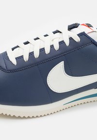 Navy blue leather sneaker with white accents, featuring a prominent white Nike logo, textured sole, and white laces with a light blue stripe.