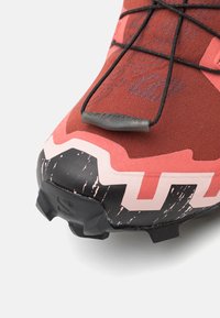 Încălțăminte sport roșie și roz, cu o parte superioară din țesătură texturată, o talpă exterioară neagră, modelată, cu crampoane distincte pentru aderentă, și vârf întărit.
