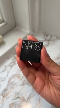 Black rectangular compact held in a hand, featuring the white embossed text "NARS" on top. Smooth surface and edges, minimalistic design.