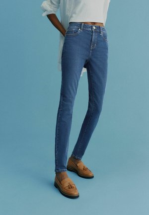 Person klädd i slim-fit blå jeans, med rullade vita ärmar och bruna tofs-loafers mot en enhetlig ljusblå bakgrund.