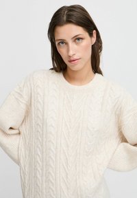 Pull en maille torsadée crème avec un col rond et des poignets côtelés. Présente des motifs texturés et une coupe ample pour le confort.