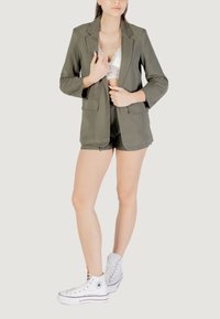 Blazer verde oliva con chiusura a un bottone, abbinato a pantaloni corti coordinati e sneaker bianche alte. Bralette in pizzo visibile sotto.