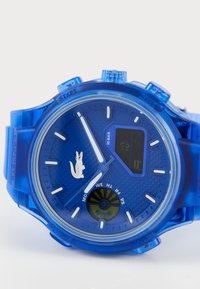 Montre numérique bleue avec un cadran texturé, des index horaires blancs et un logo de crocodile en évidence. Elle est dotée d'un bracelet en silicone transparent et de plusieurs boutons.