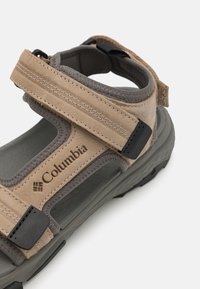 Beige semsket sandaler med grå detaljer, justerbare stropper, robust såle og Columbia-logo. Teksturerte stropper og en polstret fotseng.