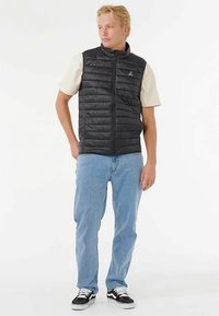Zwart gewatteerd vest met een hoge kraag, ritssluiting en horizontale stiksels. Draagt over een crèmekleurig shirt en gecombineerd met blauwe spijkerbroeken en zwarte sneakers.