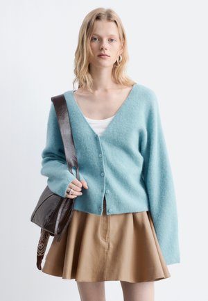 Junge Frau trägt eine hellblaue, geknöpfte Strickjacke, ein weißes Top, einen braunen Rock und trägt eine dunkelbraune Schultertasche.