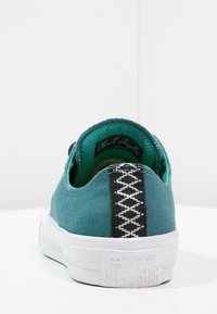 Baskets en toile bleu sarcelle avec une semelle en caoutchouc blanche, un détail tressé noir au talon et un intérieur vert contrastant. Présente le logo "Converse All Star".