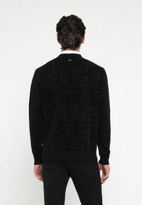 Μαύρο πλεκτό πουλόβερ με υφασμένο μοτίβο, στρογγυλή λαιμόκοψη και ριμπ μανσέτες. Η πλάτη διαθέτει διακριτικό λογότυπο στη γραμμή του λαιμού.