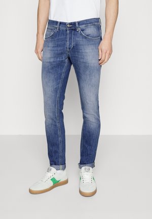 Homme portant un jean bleu délavé slim et des baskets blanches avec des accents verts, debout devant un fond clair uni.