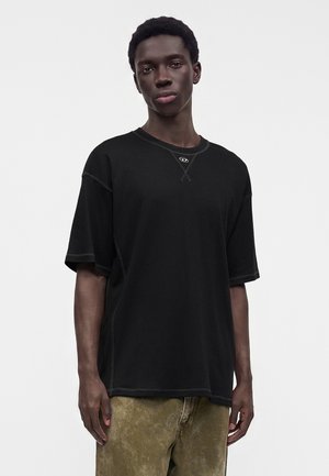 Jeune homme aux cheveux courts portant une chemise noire ample à manches courtes et un pantalon vert olive délavé sur un fond clair uni.