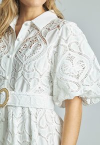 Biała sukienka koszulowa z koronkowym wykończeniem i krótkimi bufiastymi rękawami, ozdobiona skomplikowanymi wzorami, z kołnierzykiem i materiałowym paskiem z okrągłą klamrą.
