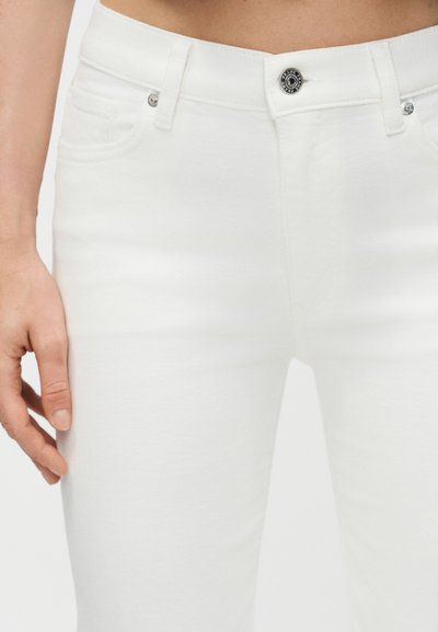 Κοντινό πλάνο ατόμου που φοράει λευκό τζιν με ψηλή μέση, μεταλλικό κουμπί και εξαρτήματα, με το αριστερό χέρι χαλαρό στο πλάι, πάνω σε λευκό φόντο.