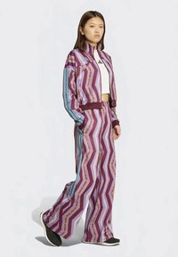 Lila und mehrfarbiger Trainingsanzug mit Zickzackmuster, bestehend aus einer Jacke mit Reißverschluss und locker sitzenden Hosen. Das Material scheint glatt und leicht zu sein.