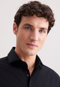Camicia nera con colletto, chiusura a bottoni, tessuto liscio, vestibilità sagomata e leggero lucido, indossata da un uomo con capelli corti e ondulati.