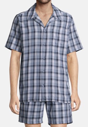 Mężczyzna ubrany w kraciastą, niebiesko-szarą koszulę na krótki rękaw zapinaną na guziki oraz pasujące kraciaste spodenki, stojący na jednolitym tle.