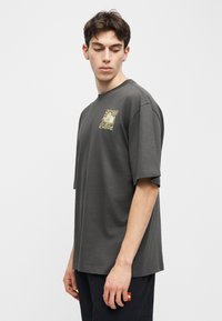 Νεαρός κοιτάζει προς τα κάτω φορώντας μια σκούρα γκρι υπερμεγέθη μπλούζα με κίτρινο γραφικό στο στήθος και μαύρο παντελόνι.