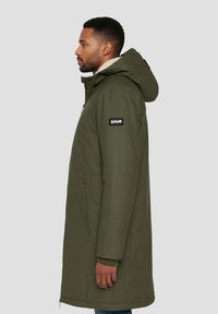 Parka larga verde oliva con capucha, cierre frontal con cremallera, bolsillos laterales y forro de Sherpa. Tiene un parche negro con logo en el hombro.