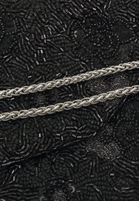 Zilveren gevlochten ketting op een gestructureerde zwarte kralenachtergrond, met verschillende kralenvormen en -groottes, die een gelaagd ontwerp creëren.