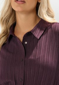 Camicia borgogna con texture, colletto, chiusura a bottoni e sottili righe verticali, realizzata in un tessuto leggero.