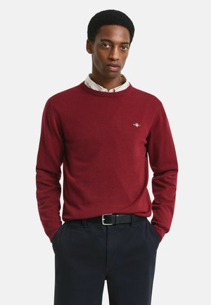 Burgunderfarbenes Sweatshirt aus weichem Stoff, mit Rundhalsausschnitt und gerippten Bündchen. Kleines Logodetail auf der linken Brust. Über einem weißen Hemd mit Kragen getragen.
