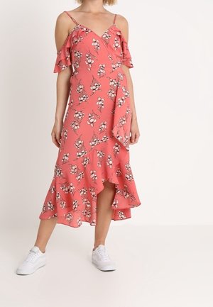 Kobieta ubrana w koralowo-różową sukienkę kopertową w kwiaty z falbanami i odkrytymi ramionami, w zestawieniu z białymi trampkami.