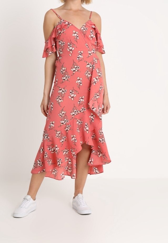 Femme portant une robe portefeuille florale rose corail avec des détails à volants et des manches épaules dénudées, associée à des baskets blanches.