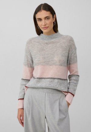 Vrouw met steil bruin haar draagt een grijs- en roze gestreepte gebreide trui en lichte grijze broek, staande tegen een effen achtergrond.