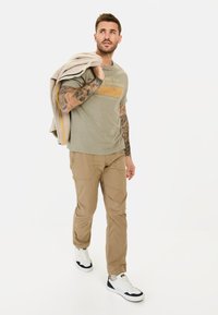 Lys grå t-shirt med orange tekst, beige cargo-bukser og hvide sneakers med sorte detaljer, bærende en let jakke over skulderen.