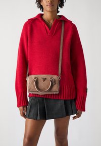 Maglione rosso a maglia con scollo a V, polsini a coste e una gonna scozzese corta. Una borsa beige con motivi in rilievo e manici doppi è indossata a tracolla.