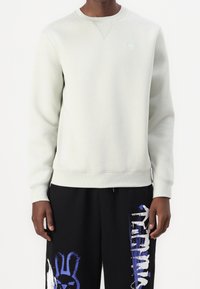 Ljusgrå sweatshirt med rundad halsringning och ribbade ärmar. Har en liten logga på bröstet, tillsammans med svarta byxor med grafiska tryck.