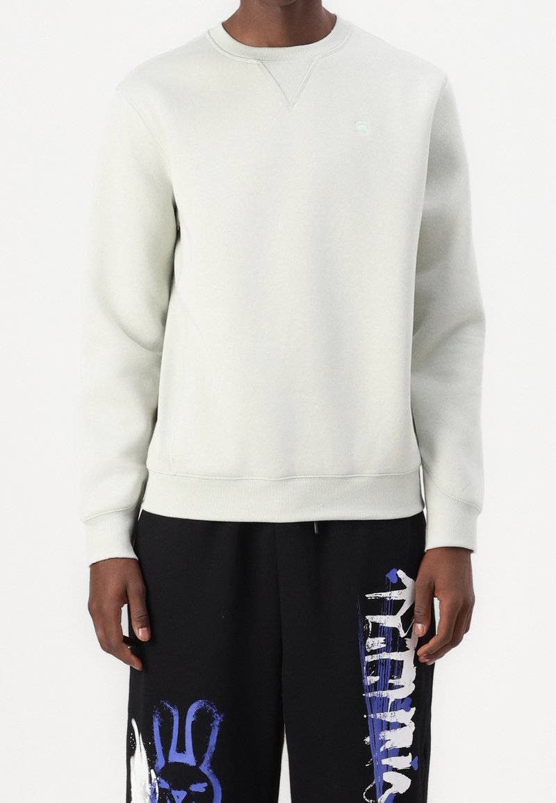 Ljusgrå sweatshirt med rundad halsringning och ribbade ärmar. Har en liten logga på bröstet, tillsammans med svarta byxor med grafiska tryck.