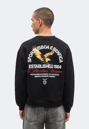 Personne aux cheveux courts portant un sweat-shirt noir avec un aigle jaune et un texte blanc et rouge faisant référence au Sport Lisboa e Benfica dans le dos.