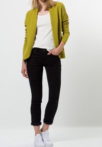 Grüner Strickcardigan, weißes T-Shirt, schwarze Jeans mit aufgerollten Bündchen und weiße Sneaker. Fokus auf die leuchtende Farbe und die strukturierte Stoffbeschaffenheit des Cardigans.