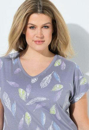 Mujer rubia con ojos azules que lleva una camiseta gris con cuello en V decorada con plumas de colores sobre un fondo claro.