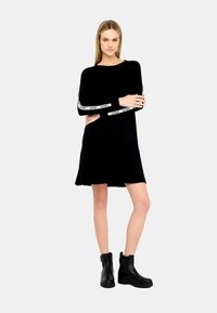 Vestito nero a maniche lunghe con gonna a ruota, dotato di accenti in bianco del logo sulle maniche. Abbinato a stivali neri fino alla caviglia. Tessuto morbido.