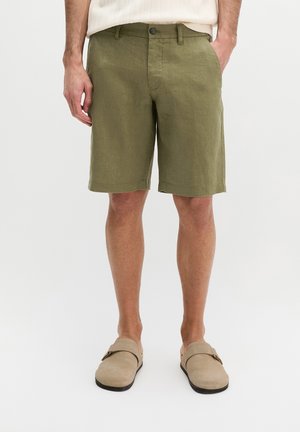 Mann trägt olivgrüne knielange Shorts, beige Slip-On-Schuhe und ein weißes Hemd, steht mit den Händen in den Taschen.