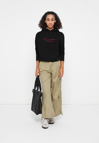 Hanorac negru cu buzunar frontal și broderie roșie, pantaloni cargo khaki cu multiple buzunare, adidași gri și o geantă tote neagră.
