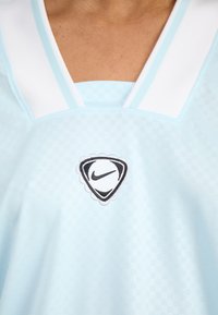 Lys blå atletisk trøje med et ternet mønster, hvid krave med detaljer, og et sort Nike-logo i en bølget kontur.