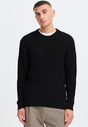 Mand iført en sort ribstrikket sweater over en hvid skjorte og beige bukser mod en ensartet lys baggrund.