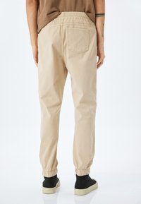 Beige katoenen joggingbroek met een elastische tailleband, aangesloten enkels en een enkele achterzak; gedragen met zwarte instap schoenen.